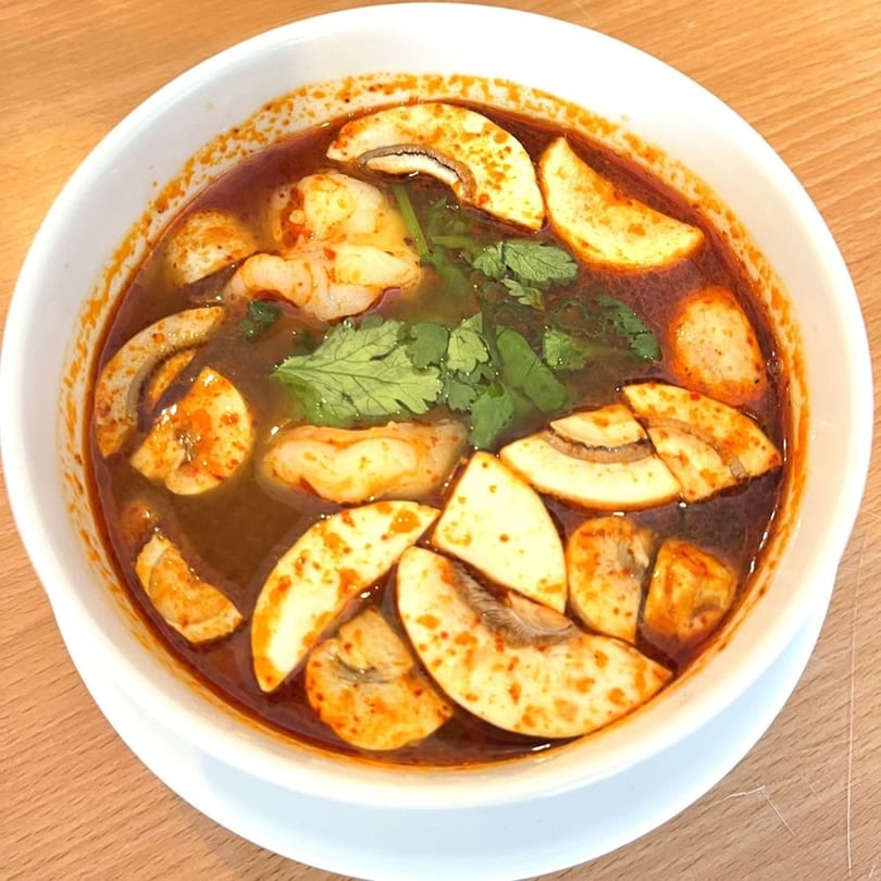 Spicy Tom Yum soup with shrimp, mushrooms, and cilantro in a white bowl on a wooden table.
