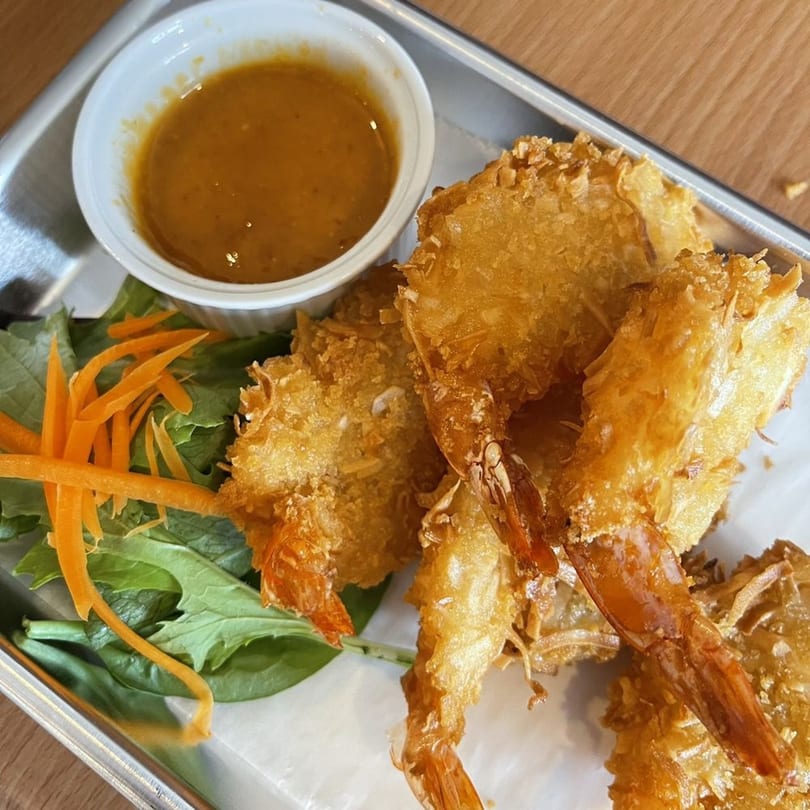 Crispy coconut shrimp with a side of peanut sauce and a garnish of greens and shredded carrots.